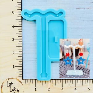 May include: A light blue silicone mold for crafting earrings, with a ruler for measurement. The image also shows a pair of star-shaped earrings with red and blue accents, displayed on a white stand. The earrings are a patriotic theme.