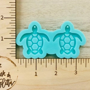 May include: Two turquoise silicone molds of sea turtles, each measuring approximately 1 inch in diameter. The molds are on a wooden ruler with inch markings.
