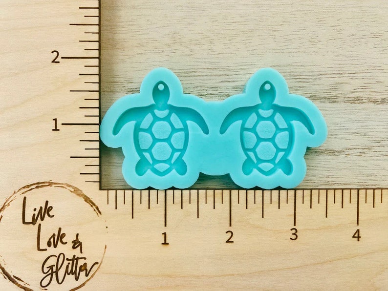 Pode incluir: Dois moldes de silicone turquesa em forma de tartarugas marinhas, cada um medindo aproximadamente 2,5 cm de di&acirc;metro. Os moldes est&atilde;o em uma r&eacute;gua de madeira com marcas em polegadas.