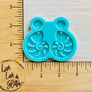 May include: A turquoise silicone mold for making earrings. The mold features two seashell designs, each with a spiral pattern and a small shell at the top. The mold is on a wooden surface with a ruler for measurement.