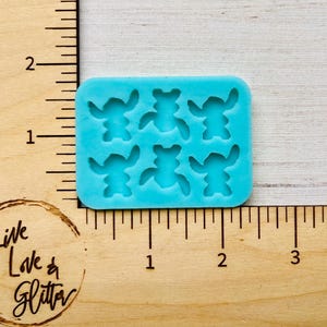 Puede incluir: Un molde de silicona azul claro con seis formas de personajes de dibujos animados. El molde es rectangular con esquinas redondeadas y est&aacute; sobre una superficie de madera con medidas en cent&iacute;metros. El texto "Live Love & Glitter" es visible.