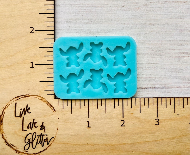 Puede incluir: Un molde de silicona azul claro con seis formas de personajes de dibujos animados. El molde es rectangular con esquinas redondeadas y est&aacute; sobre una superficie de madera con medidas en cent&iacute;metros. El texto "Live Love & Glitter" es visible.