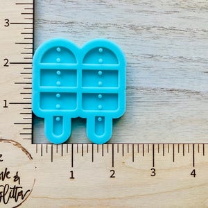 May include: A blue silicone mold shaped like two popsicles, each with four sections and a stick. The mold is about 2 inches tall and is shown next to a ruler.