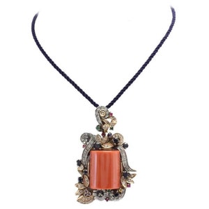 Puede incluir: Un collar con colgante que presenta una piedra rectangular de color coral engastada en un marco ornamentado. El marco está adornado con diamantes, zafiros y esmeraldas, y está suspendido de un cordón azul oscuro.