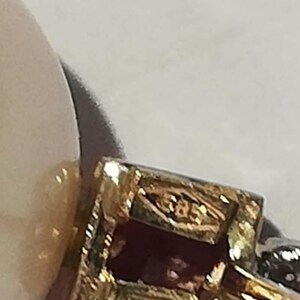 May include: Close-up of a gold-coloured jewellery piece with a rectangular design and a red stone. The jewellery has intricate detailing and a small marking that reads "585". The background is a light colour.
