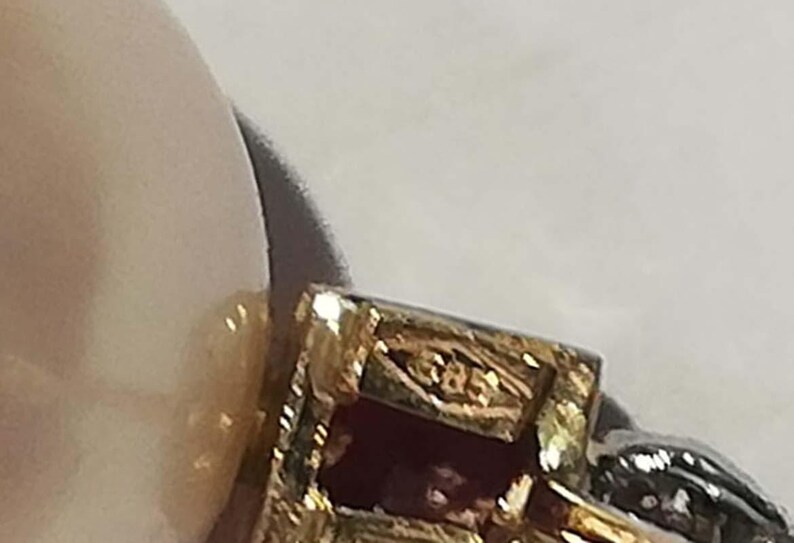 May include: Close-up of a gold-coloured jewellery piece with a rectangular design and a red stone. The jewellery has intricate detailing and a small marking that reads "585". The background is a light colour.