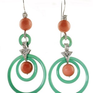 May include: A pair of Art Deco-style earrings. Each earring features a coral-colored bead, a green hoop, and a diamond-studded accent. The earrings have a silver hook and a second coral-colored bead at the bottom, with a green hoop that spirals around the bead.