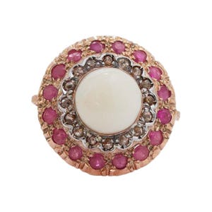 May include: A vintage-style ring with a central off-white stone, surrounded by a halo of small, vibrant red gemstones. The ring's setting is a warm rose gold color, with intricate detailing around the stones.
