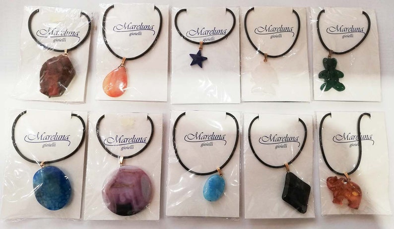 May include: A collection of necklaces with various pendant shapes, including a star, bear, and elephant. Each necklace features a black cord and a gold-colored clasp. The pendants are made of different materials and colors, and are displayed on white cards with the "Mareluna gioielli" logo.