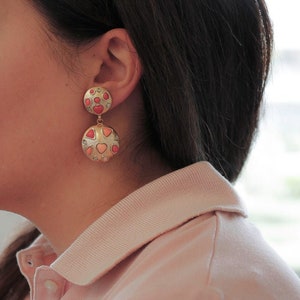 May include: Gold dangle earrings with two round discs. The discs are covered in small pink, red, and white hearts.