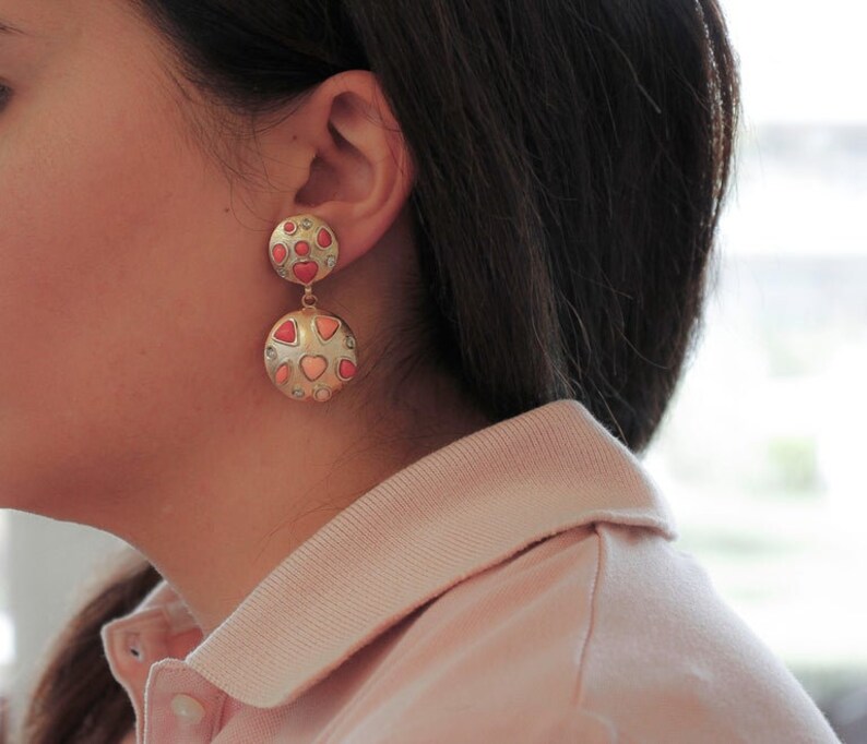 May include: Gold dangle earrings with two round discs. The discs are covered in small pink, red, and white hearts.