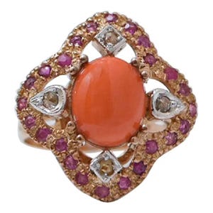May include: A gold ring featuring a central oval orange cabochon stone, surrounded by a decorative frame set with small round pink stones and four accent stones in silver-toned settings at the cardinal points of the oval center.