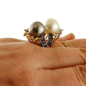 May include: A gold ring featuring a black and white pearl setting. The ring is decorated with a row of blue sapphires.