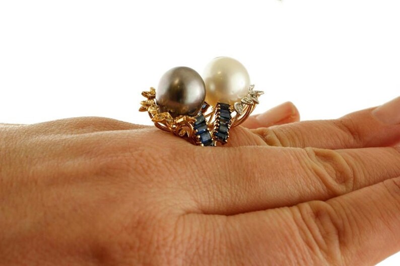 May include: A gold ring featuring a black and white pearl setting. The ring is decorated with a row of blue sapphires.