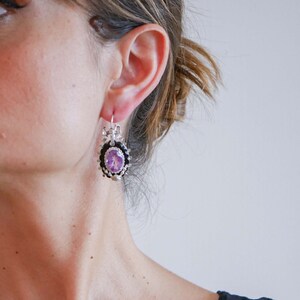May include: Silver-toned drop earrings with an oval, light purple gemstone center. The gemstone is surrounded by a black border and a decorative silver frame. The earrings have a floral design and are suspended from a silver hook.