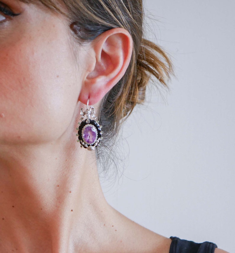 May include: Silver-toned drop earrings with an oval, light purple gemstone center. The gemstone is surrounded by a black border and a decorative silver frame. The earrings have a floral design and are suspended from a silver hook.