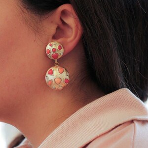 May include: A pair of gold dangle earrings with pink and white heart-shaped stones. The earrings are made of metal and have a polished finish.