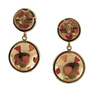 May include: Gold tone dangle earrings with two round discs. The larger disc has a textured surface with pink and red enamel hearts and geometric shapes. The smaller disc has a similar design with a red enamel heart.