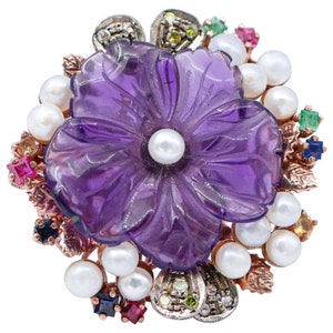 May include: A purple amethyst flower brooch with a carved design, surrounded by pearls and colorful gemstones.