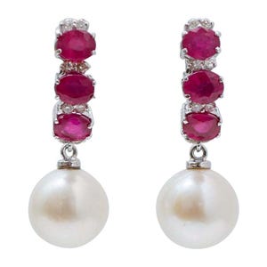 Puede incluir: Un par de pendientes elegantes con tres rubíes rojos ovalados y pequeños diamantes engastados en metal plateado. Cada pendiente tiene una gran perla blanca redonda colgando debajo. Fondo blanco.