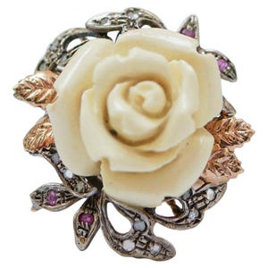 May include: A cream-colored rose ring with detailed petals, surrounded by silver and gold-toned leaves. The ring features small pink and clear gemstones, creating a floral design. The ring is set against a white background.