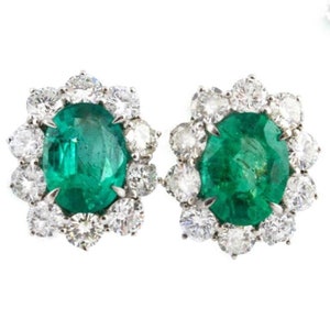 May include: A pair of stud earrings featuring oval-cut emeralds surrounded by a halo of round-cut diamonds. The emeralds are a vibrant green, and the diamonds sparkle against a white background. The earrings are set in a silver metal.