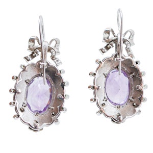 May include: A pair of silver earrings with an oval, faceted, light purple gemstone center. The earrings have a scalloped edge and a bow detail at the top. Small, round, clear stones are set around the oval gemstone.