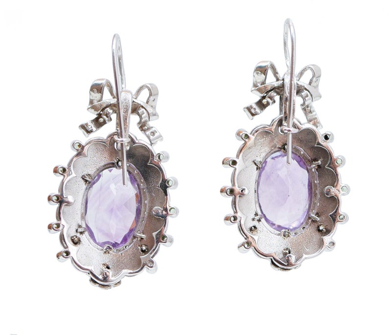 May include: A pair of silver earrings with an oval, faceted, light purple gemstone center. The earrings have a scalloped edge and a bow detail at the top. Small, round, clear stones are set around the oval gemstone.