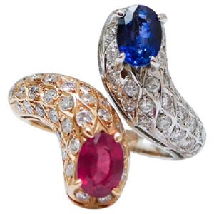 Puede incluir: Dos anillos de piedras preciosas. Uno presenta un rubí engastado en oro, rodeado de diamantes. El otro tiene un zafiro azul engastado en plata, también rodeado de diamantes. Ambos anillos tienen un diseño curvo.