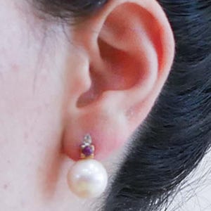May include: Close-up of a pearl earring. The earring features a large, round, white pearl suspended below a small gold setting with a red gemstone and a small clear stone. The earring is attached to an ear.