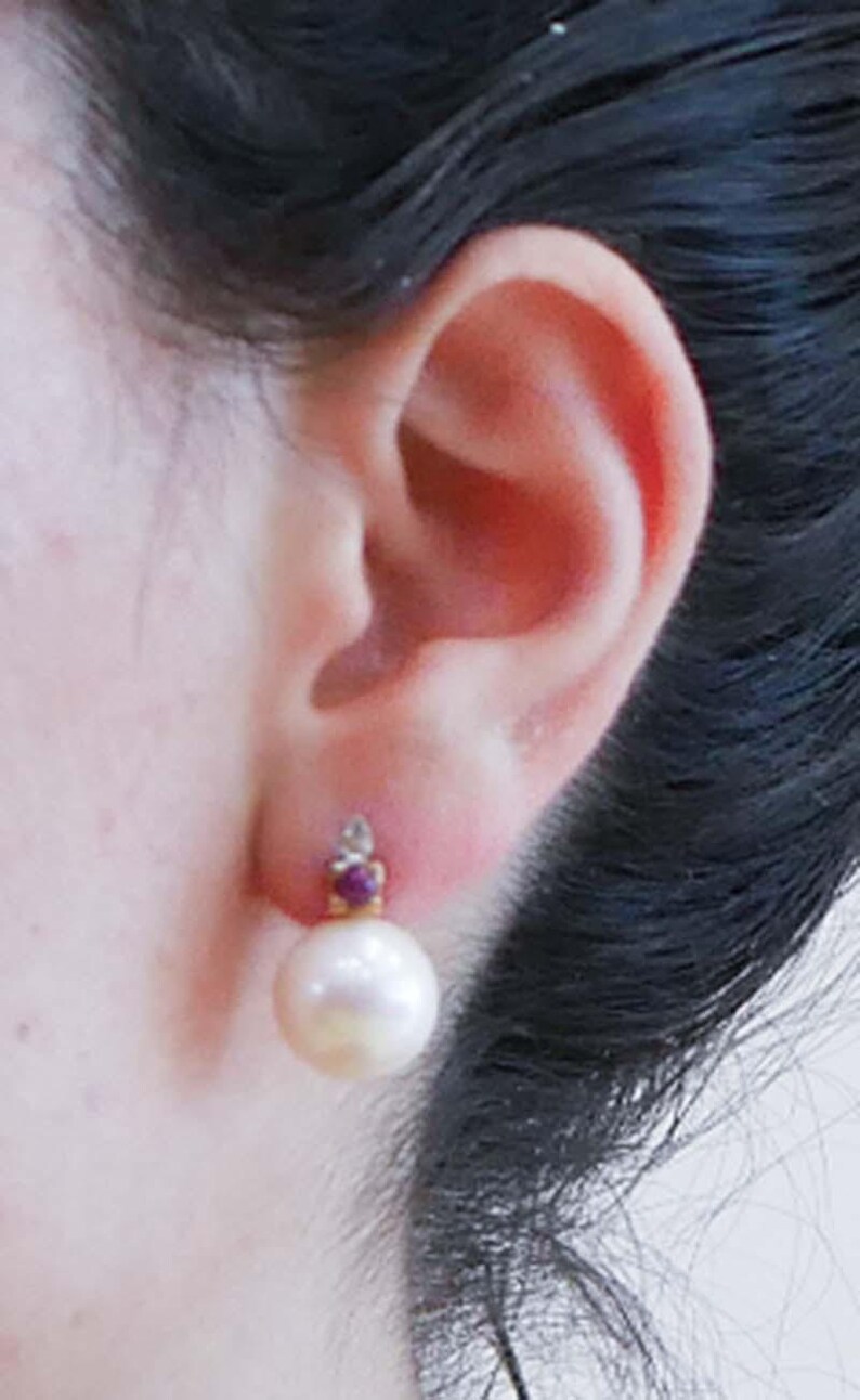 May include: Close-up of a pearl earring. The earring features a large, round, white pearl suspended below a small gold setting with a red gemstone and a small clear stone. The earring is attached to an ear.
