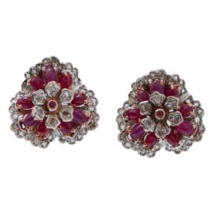 May include: A pair of floral-shaped earrings featuring oval-cut deep red rubies arranged in a cluster, surrounded by small, clear, round-cut diamonds set in a metallic frame, creating a layered, multi-petaled appearance with a central ruby.