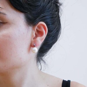 May include: A close-up of a pearl earring with a small red gemstone above it. The pearl is large and round, with a creamy white colour. The earring is set against the backdrop of a person's ear and dark hair.