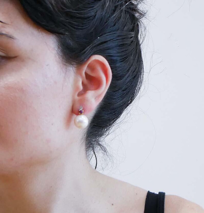 May include: A close-up of a pearl earring with a small red gemstone above it. The pearl is large and round, with a creamy white colour. The earring is set against the backdrop of a person's ear and dark hair.