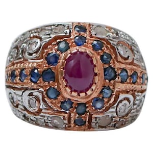 Op de afbeelding: Een brede ring met een centrale ovale cabochon robijn gezet in roségoud, omringd door een halo van kleine ronde blauwe saffieren. De zilverkleurige metalen basis is getextureerd met ingewikkeld krulwerk en geaccentueerd met kleine heldere stenen.