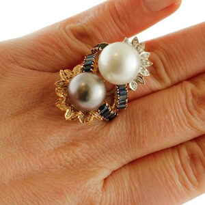 May include: A gold ring featuring two pearls, one white and one black, set in a floral design with blue sapphires.