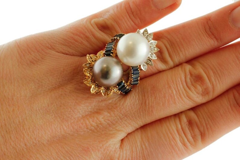 May include: A gold ring featuring two pearls, one white and one black, set in a floral design with blue sapphires.