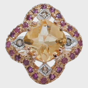 Op de afbeelding: Een ring met een centrale gele edelsteen in kussenslijpvorm, omringd door een geschulpte rand van kleine ronde roze stenen. De zetting van roségoud bevat vier zilverkleurige ruitvormige accenten, elk met een kleine heldere steen.
