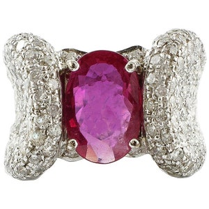May include: A silver ring featuring a large oval ruby gemstone at the center. The ring's design includes two bow-shaped sections encrusted with small, sparkling diamonds on either side of the ruby.