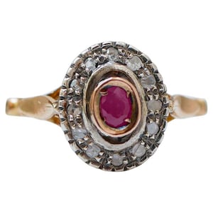 Pode incluir: Anel de rubi oval com um halo de pequenos diamantes. O anel apresenta uma banda de ouro e uma moldura de prata em torno do rubi e dos diamantes. O rubi é de cor vermelha profunda e os diamantes brilham.