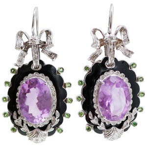 May include: A pair of ornate earrings featuring oval amethyst gemstones set in black enamel frames. The earrings have a scalloped edge, accented with green gemstones and a bow design at the top. The earrings are made of silver.