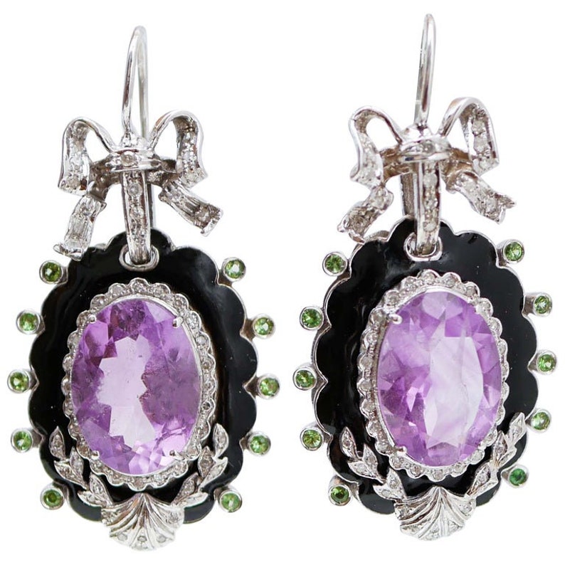 May include: A pair of ornate earrings featuring oval amethyst gemstones set in black enamel frames. The earrings have a scalloped edge, accented with green gemstones and a bow design at the top. The earrings are made of silver.