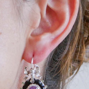 May include: A silver earring with a black and silver frame, featuring a purple gemstone center. The earring has a bow and leaf design at the top and is accented with green stones around the black frame. The earring is hanging from a silver hook.