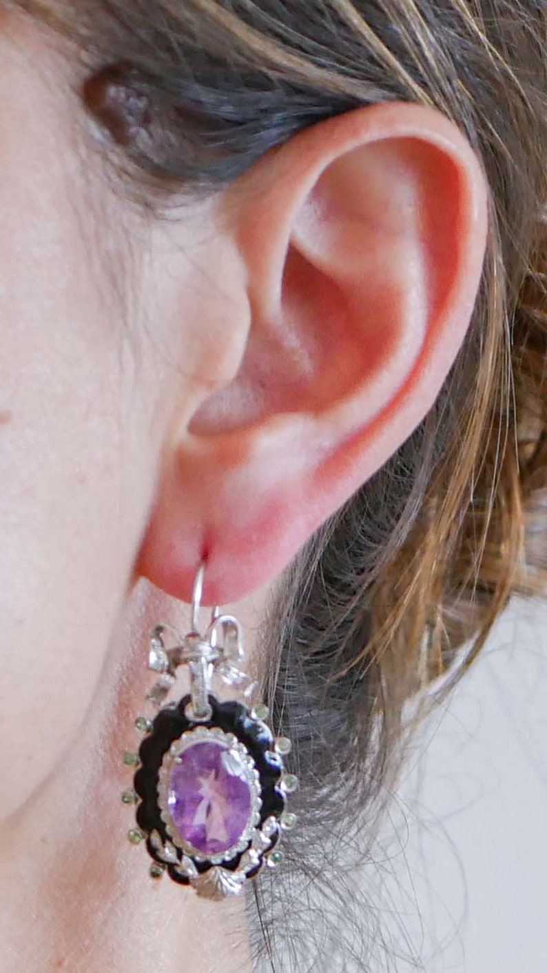 May include: A silver earring with a black and silver frame, featuring a purple gemstone center. The earring has a bow and leaf design at the top and is accented with green stones around the black frame. The earring is hanging from a silver hook.