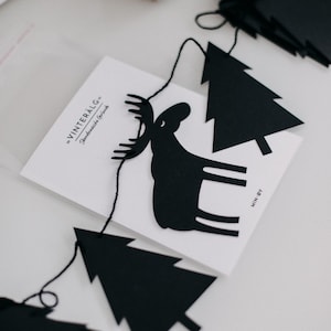 May include: Black paper garland with moose and Christmas tree cutouts. The garland includes a white card with the text "VINTERALG - Skandinavische Girlande". The garland is strung on black thread.