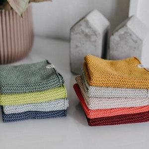 Puede incluir: Una pila de seis paños de cocina de punto en varios colores, incluyendo verde, amarillo, beige, gris y rojo. Los paños de cocina están doblados y apilados sobre una superficie blanca.
