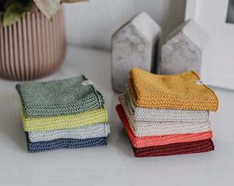 May include: A stack of six knitted dishcloths in various colors, including green, yellow, beige, gray, and red. The dishcloths are folded and stacked on a white surface.