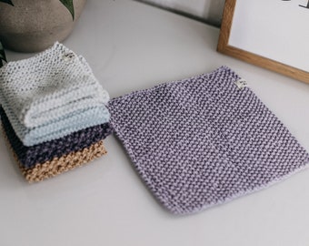 May include: A stack of four knitted dishcloths in various colors, including white, light blue, dark purple, and tan. The dishcloths are folded and stacked on a white surface. A single purple knitted dishcloth is lying flat on the surface to the right of the stack.