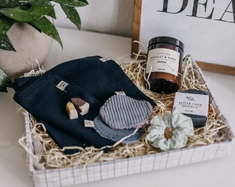 May include: A gift basket filled with various self-care items. Includes a candle labeled "Muscat & Sage", a bar of soap, dark blue washcloths, a scrunchie, and other small items. The basket is lined with straw.