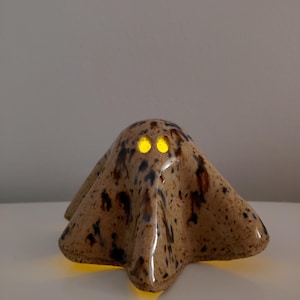 May include: A ceramic ghost figurine with a glossy, speckled tan and dark brown glaze. The figure features two hollow, circular eye openings that emit a warm, bright yellow light from within, casting a glow onto the surface below the base.